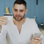 Man in a white shirt holds a credit card up while looking at his smartphone in a blue-kitchen setting, suggesting online shopping or payment.