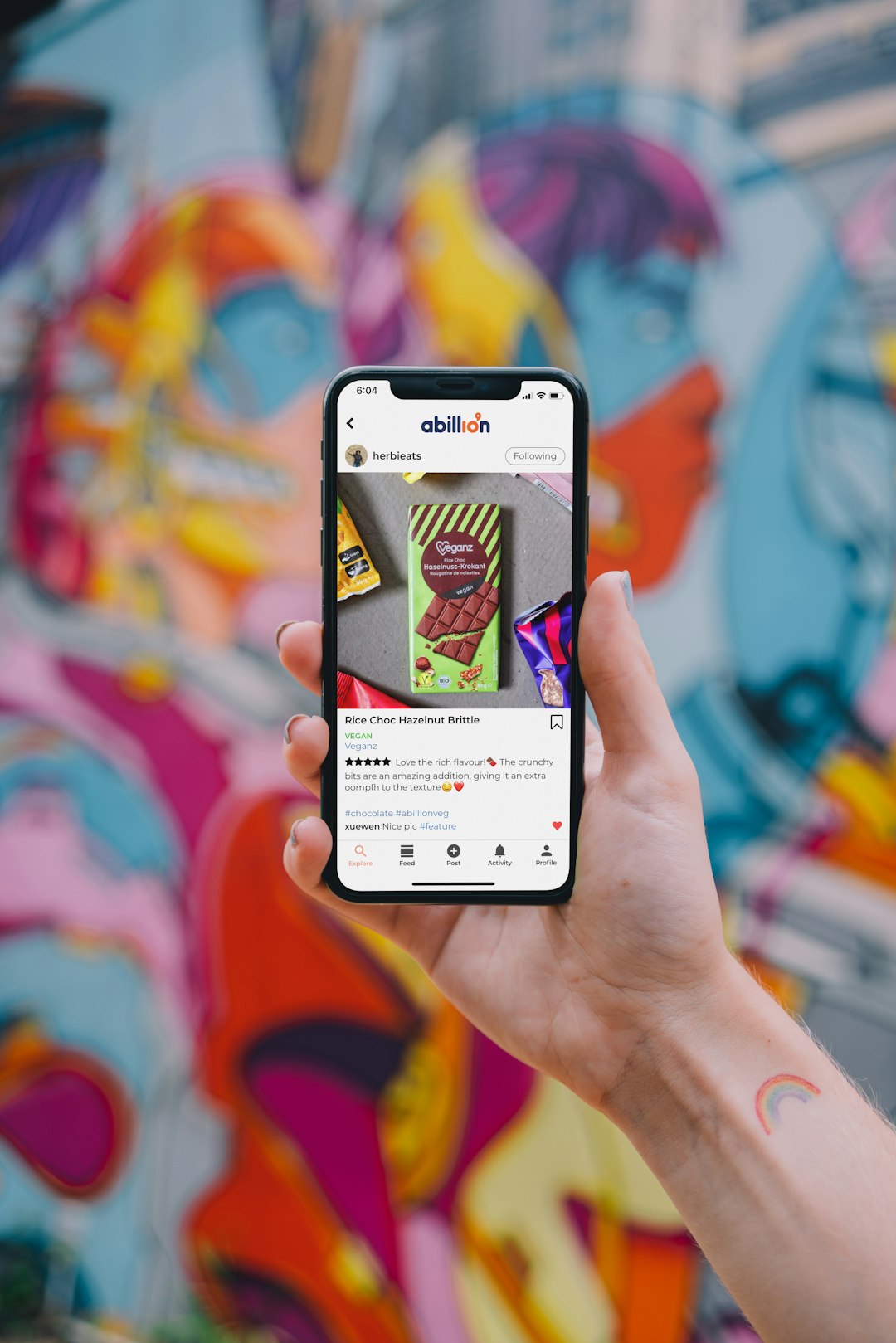 Hand steadying a smartphone showing a vegan chocolate product post against a bright graffiti wall in the background, onlookers blurred out of focus.