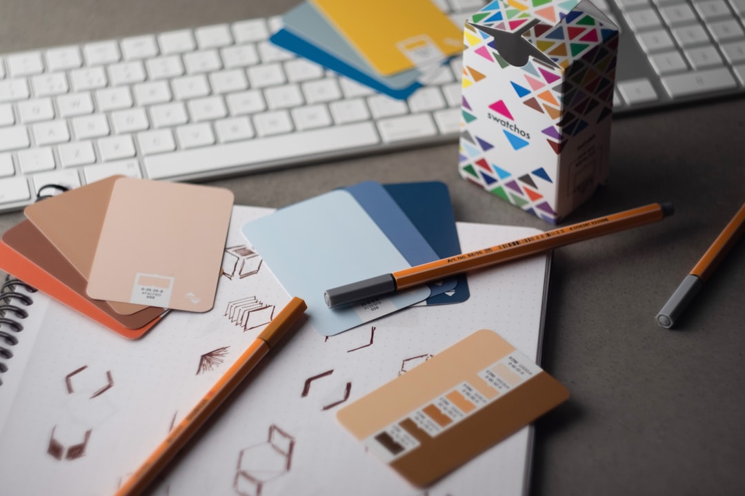 Color swatch cards and pencils arranged on an open notebook, with a colorful Swatchos box and a keyboard in the background on a desk.