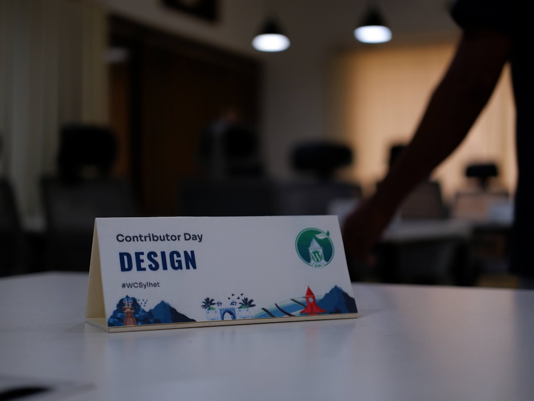 White tent card on a table reads 'Contributor Day DESIGN' with a green circular logo and a decorative blue landscape at the bottom, in a dim conference room setting.