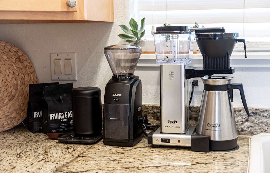 Coffee station on a granite counter: black espresso grinder, small scale, Irving Farm coffee bags, a silver Moccamaster-style drip brewer, and a stainless thermal carafe on a warming base with a plant in the background.