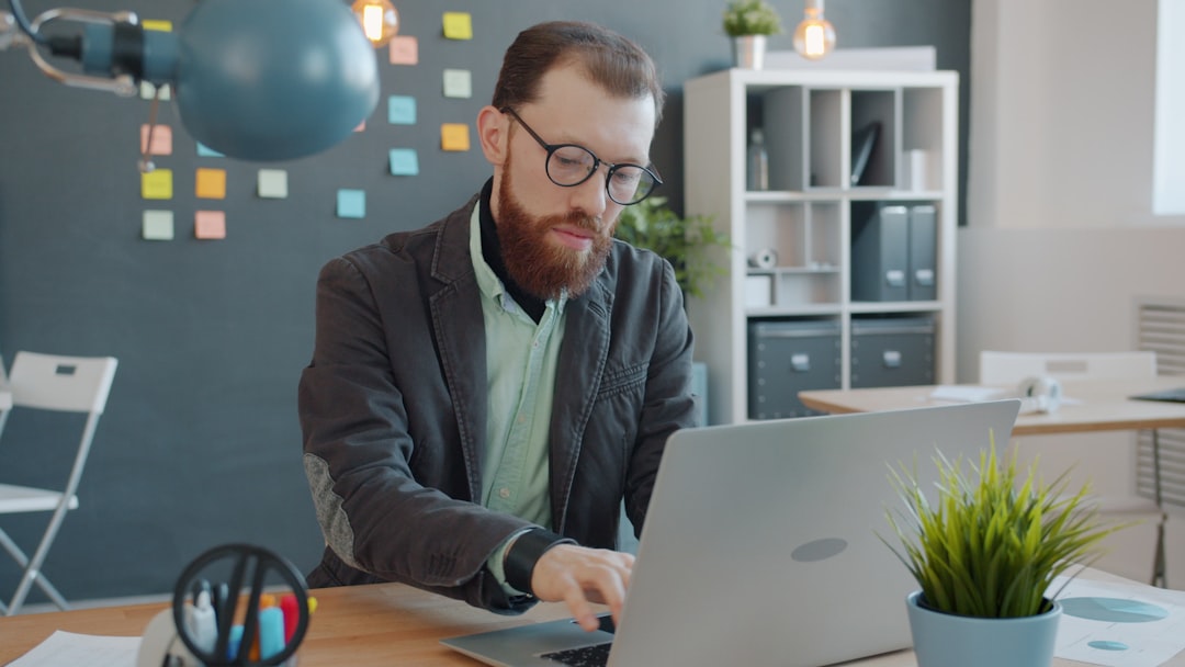 Bearded man wearing glasses typing on a silver laptop at a wooden desk in a bright, modern office','Man with a beard and glasses focused on a laptop at a tidy workspace with plants.