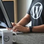 Person wearing a WordPress shirt types on a keyboard at a desk with a monitor nearby; a WordPress mug sits in the foreground.