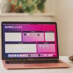 Rose gold MacBook Air open on a white desk, displaying a pink desktop/dashboard with apps, blurred green plants in the background.