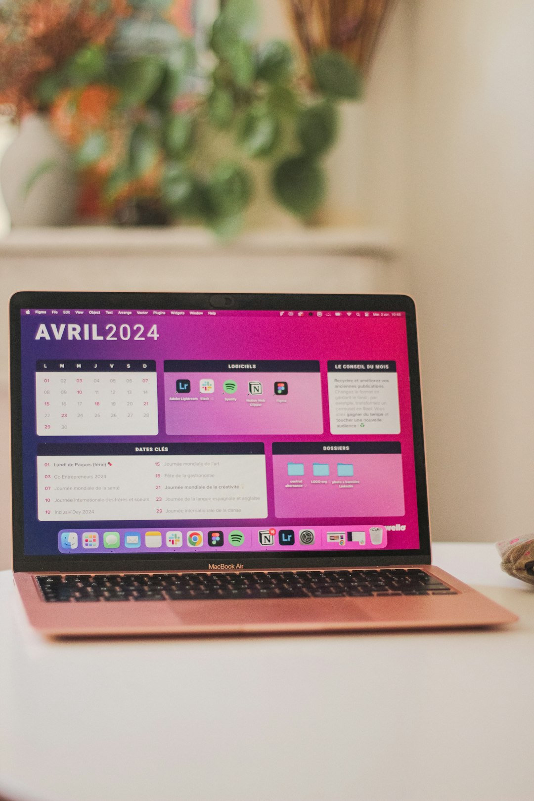 Rose gold MacBook Air open on a white desk, displaying a pink desktop/dashboard with apps, blurred green plants in the background.