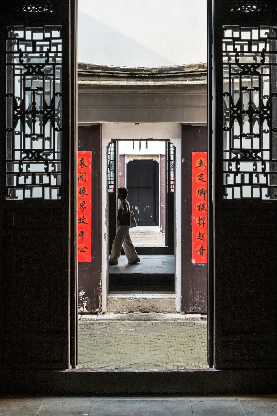 Person walks through a series of doorways in a traditional building, framed by ornate black lattice doors and red vertical banners with Chinese characters.