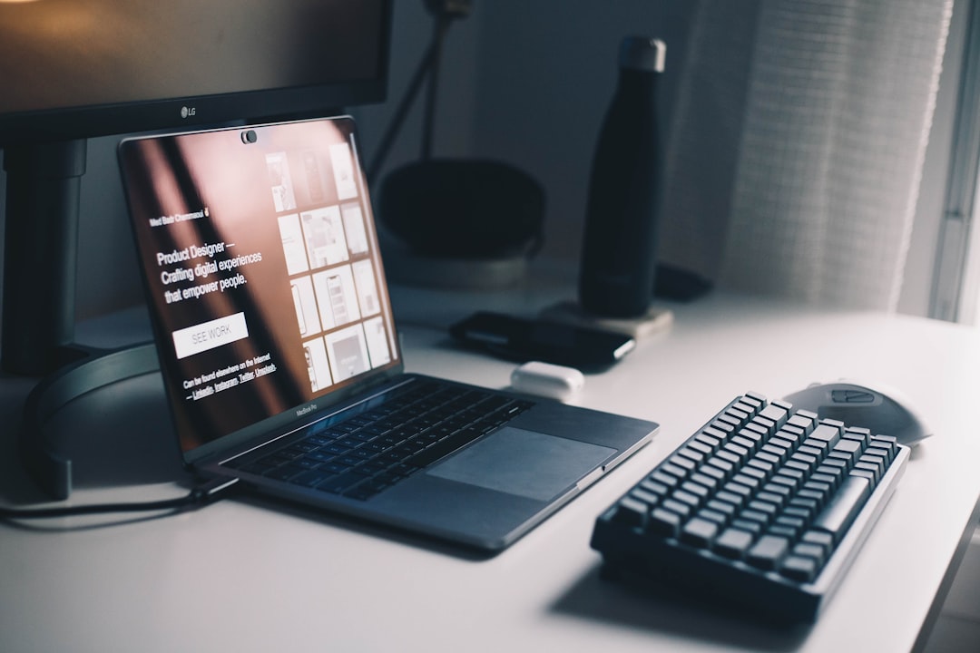 Open laptop on a white desk displaying a design portfolio, with a keyboard and mouse in the foreground and a monitor in the background.