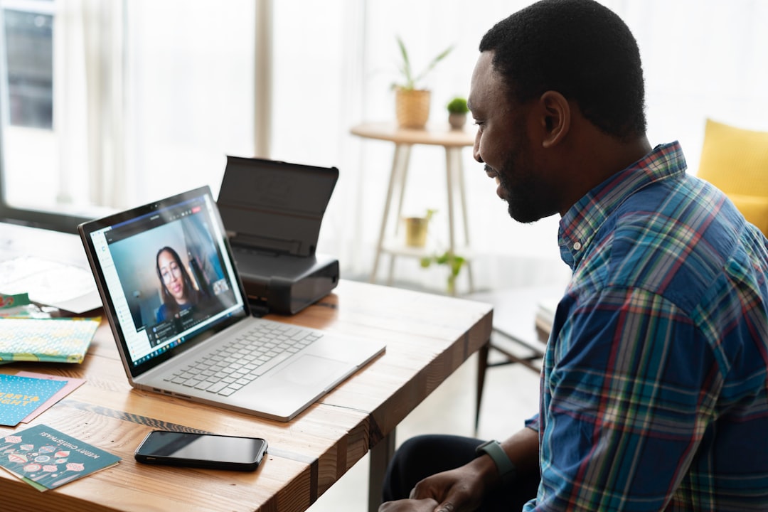 Man in a blue plaid shirt sits at a wooden desk, participating in a video call on a silver laptop.