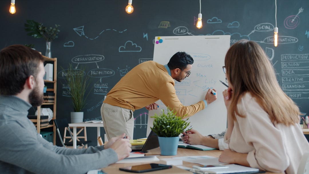 Team of three colleagues collaborating around a whiteboard in a modern office; a man in mustard sweater writes while two watch.