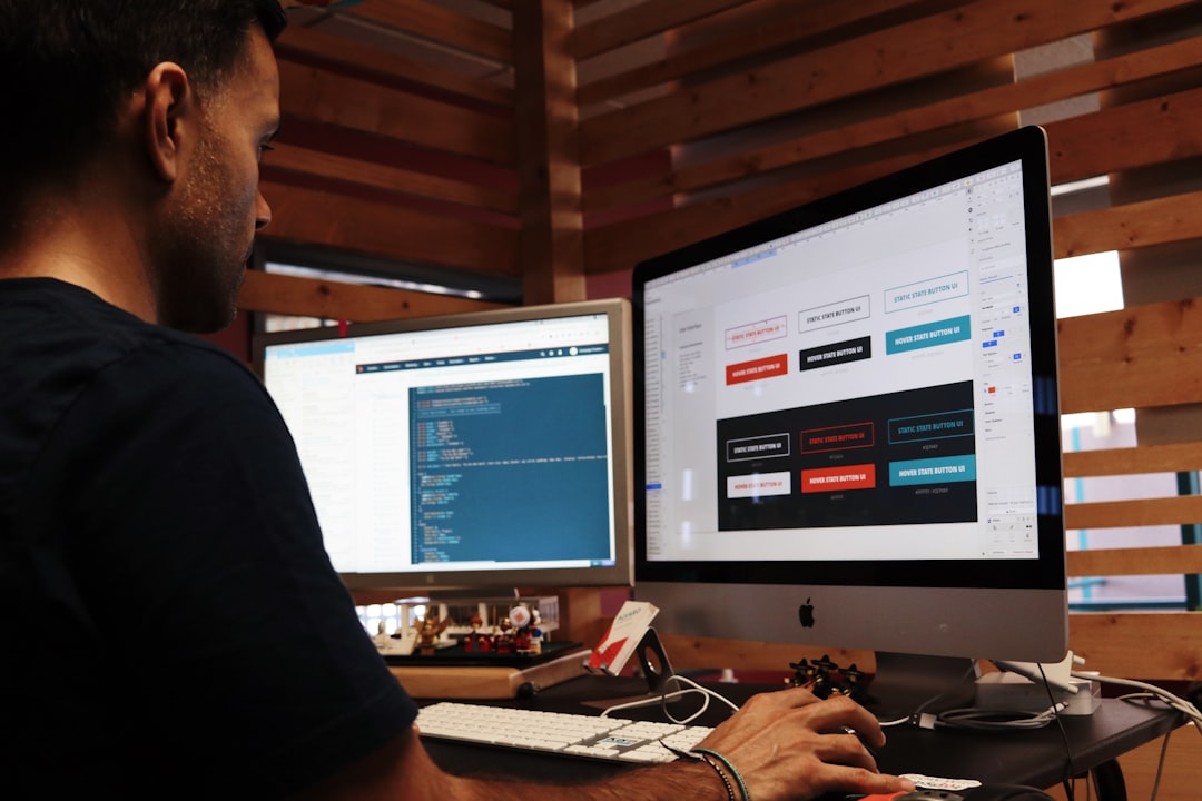 A person working at a desk with two monitor screens, one displaying code and the other a UI button design layout.