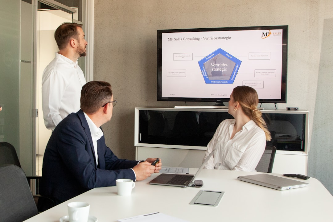 Three professionals in a modern office watching a presentation on a wall monitor.