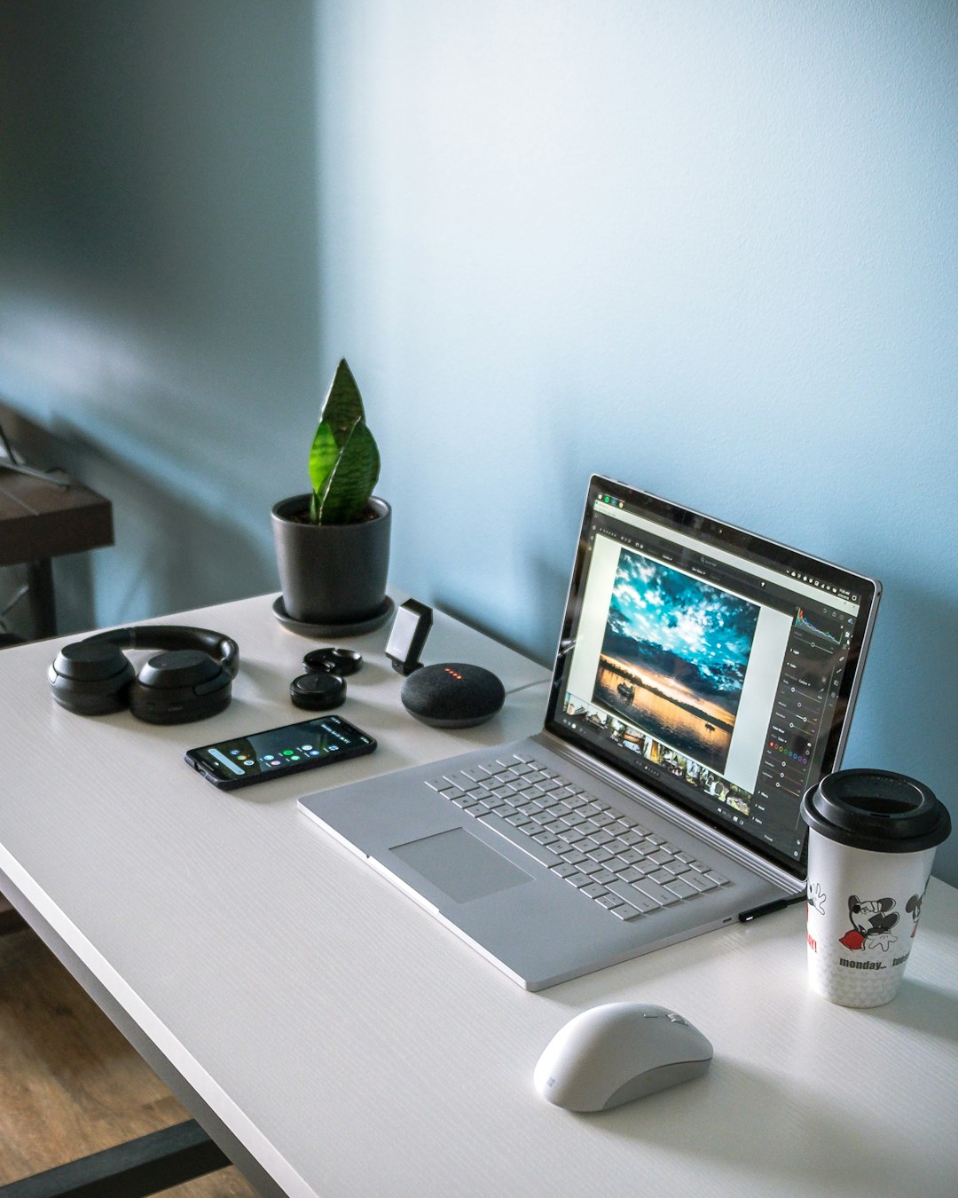 Open laptop on a white desk editing a beach photo, with a wireless mouse, coffee mug, plant, headphones, and phone nearby against a blue wall.