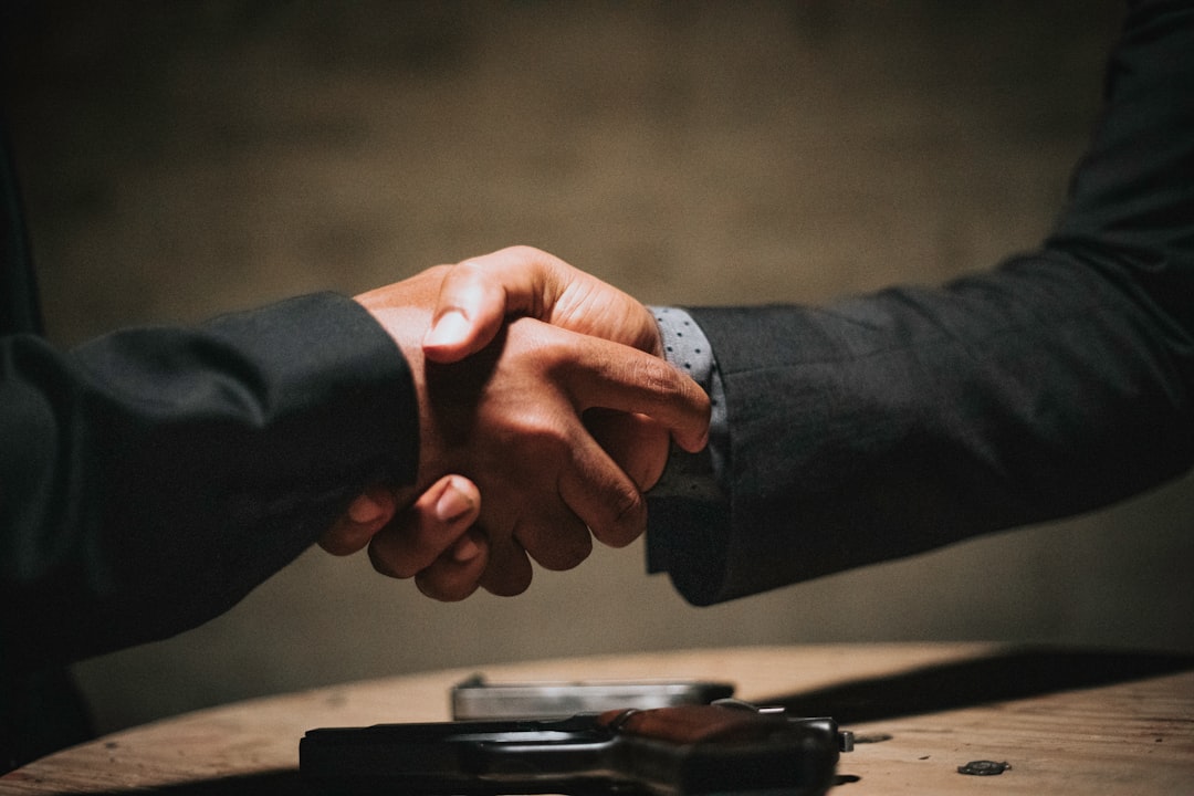 Two people in dark suits shake hands over a handgun resting on a wooden table.