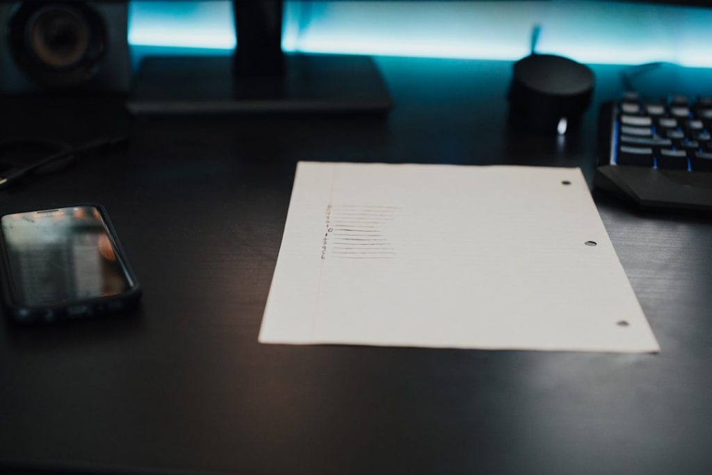 Desk workspace featuring a lined sheet of paper on a dark surface, with a smartphone to the left and a keyboard to the right under blue ambient light.