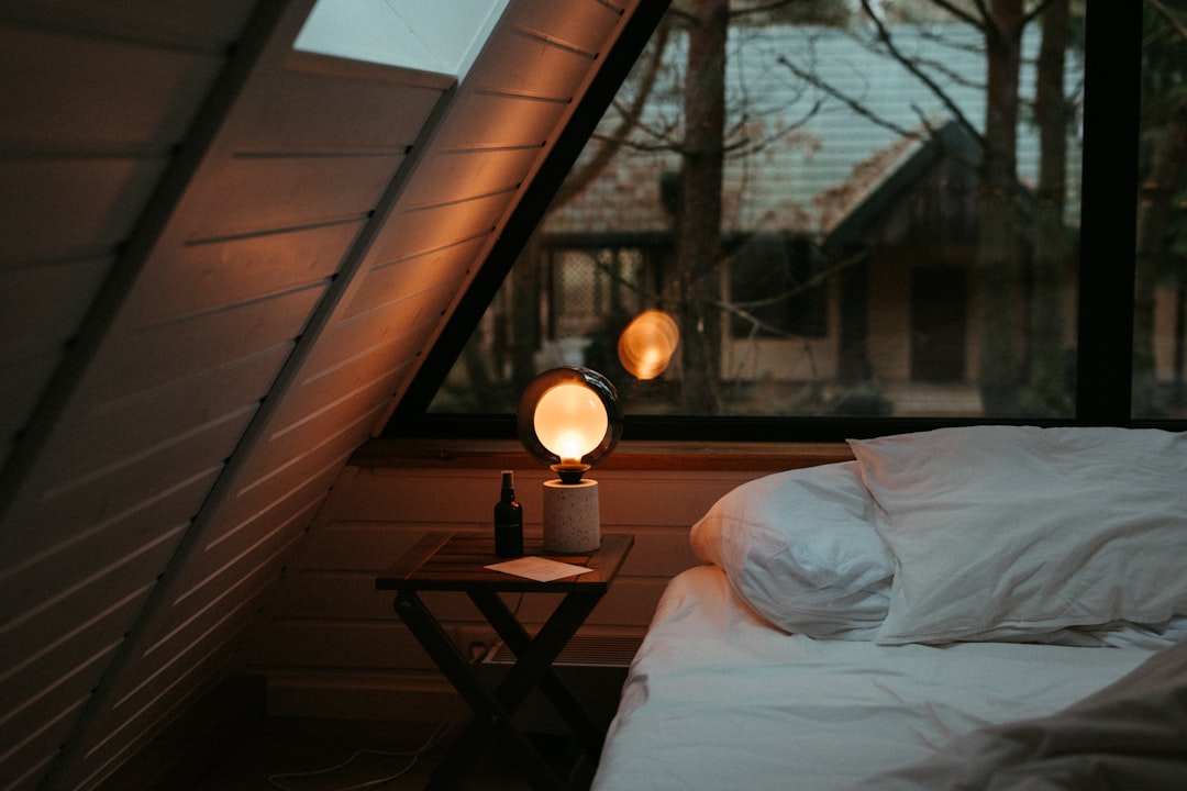 Cozy attic bedroom with a bed and white linens, a small table with a glowing round lamp and bottle beside it by a large window.