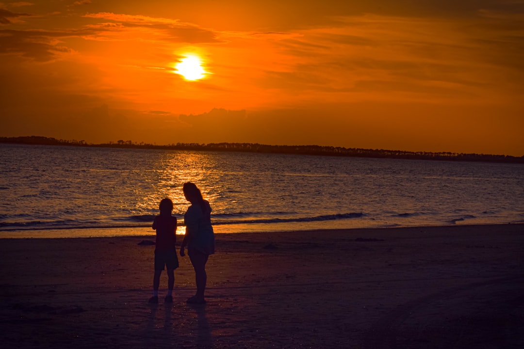 Silhouetted adults on a beach at sunset with an orange sky and reflecting water behind them.