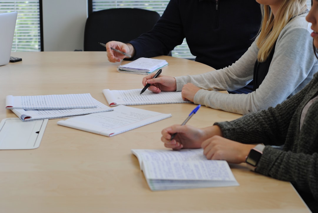 People seated around a table taking notes in notebooks; a laptop is on the left side of the table.