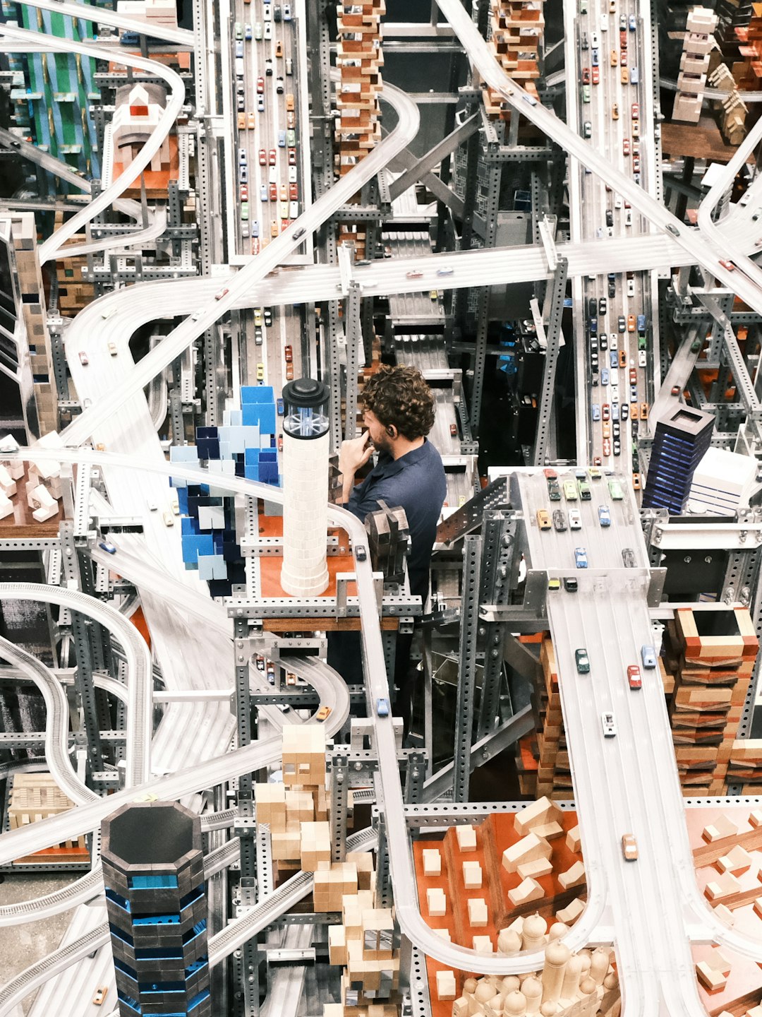 Man in a blue shirt stands among an intricate, multi-level toy track system with looping white ramps, metal supports, and tiny cars and blocks.