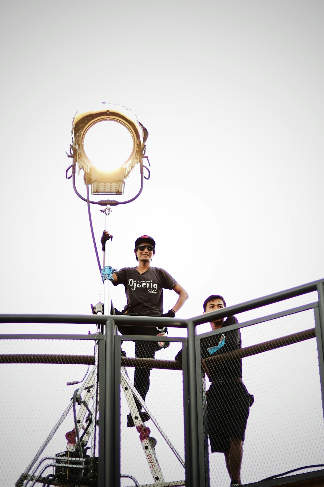 Two crew members on a raised catwalk adjust a large circular stage light mounted on a stand to prepare for filming/lighting.