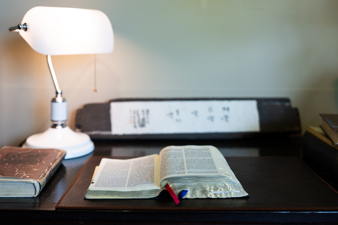 Open Bible with red and blue ribbons on a dark wooden desk beside a white desk lamp in warm light.