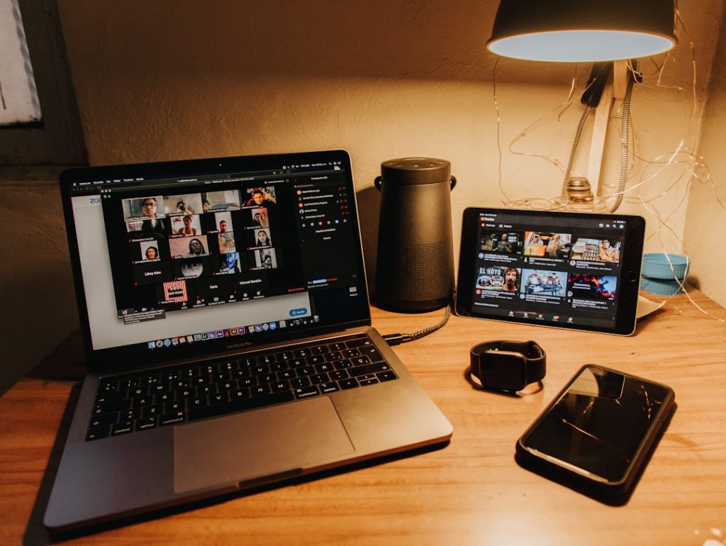 Desk setup with a laptop showing a video conference grid, a tablet playing videos, a smartwatch, a phone, and a desk lamp.
