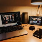 Desk setup with a laptop showing a video conference grid, a tablet playing videos, a smartwatch, a phone, and a desk lamp.