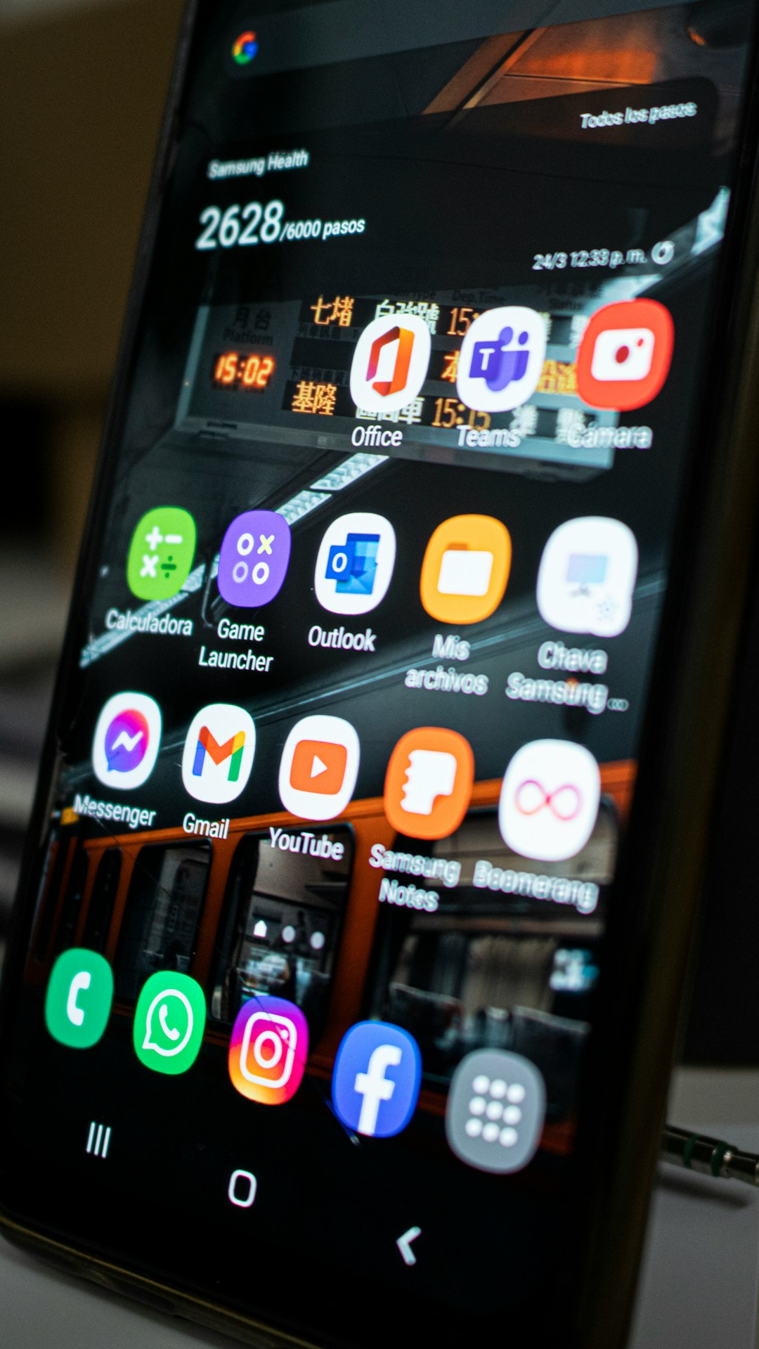 Close-up of a smartphone home screen showing app icons like Office, Teams, Outlook, Gmail, YouTube, Instagram and Messenger on a tilted display.