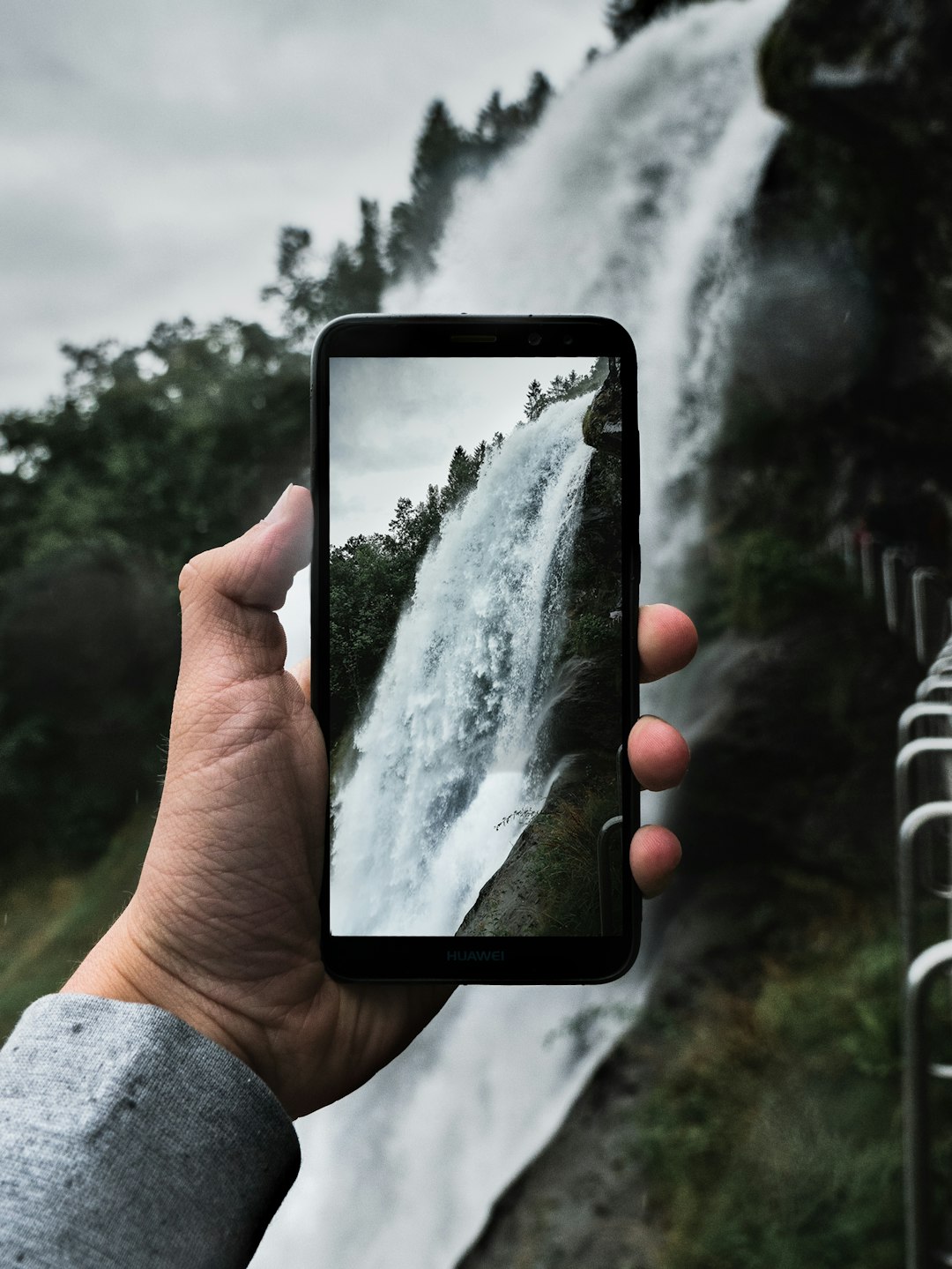 Person's hand holding a Huawei smartphone as it captures a tall waterfall against a forested backdrop and a railing on the right.