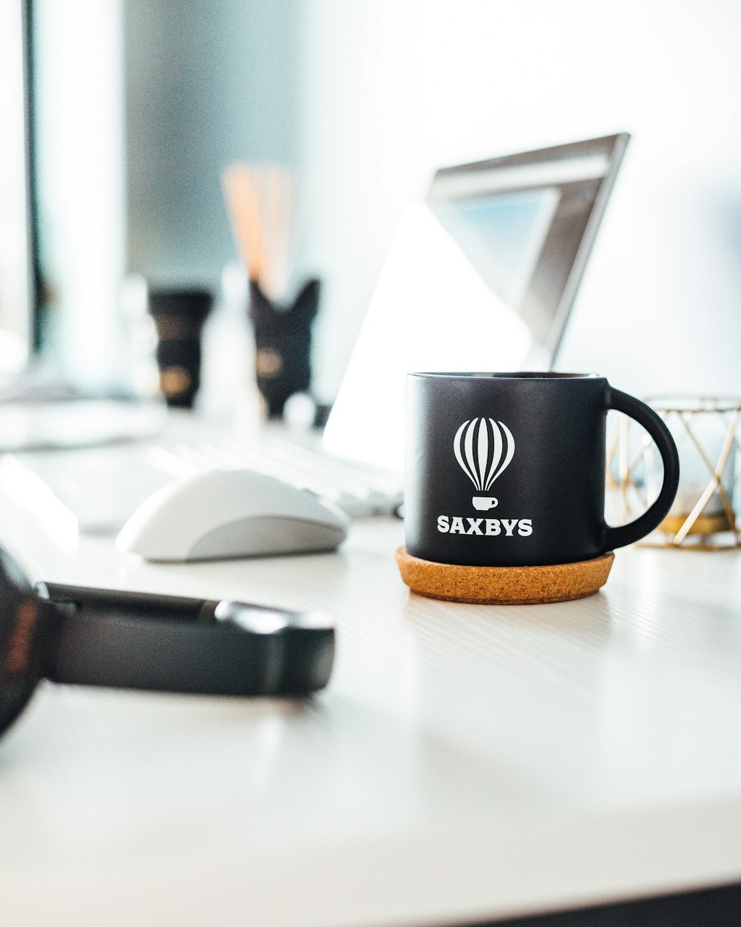 Black mug with SAXBYS logo and cork coaster on a white desk, with a laptop and keyboard in soft focus behind it.