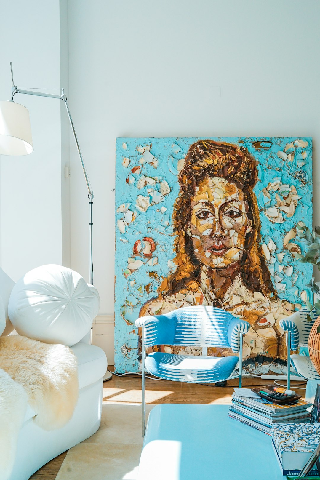Bright living room with a large mosaic portrait leaning against a turquoise wall, flanked by a blue chair and white sofa.
