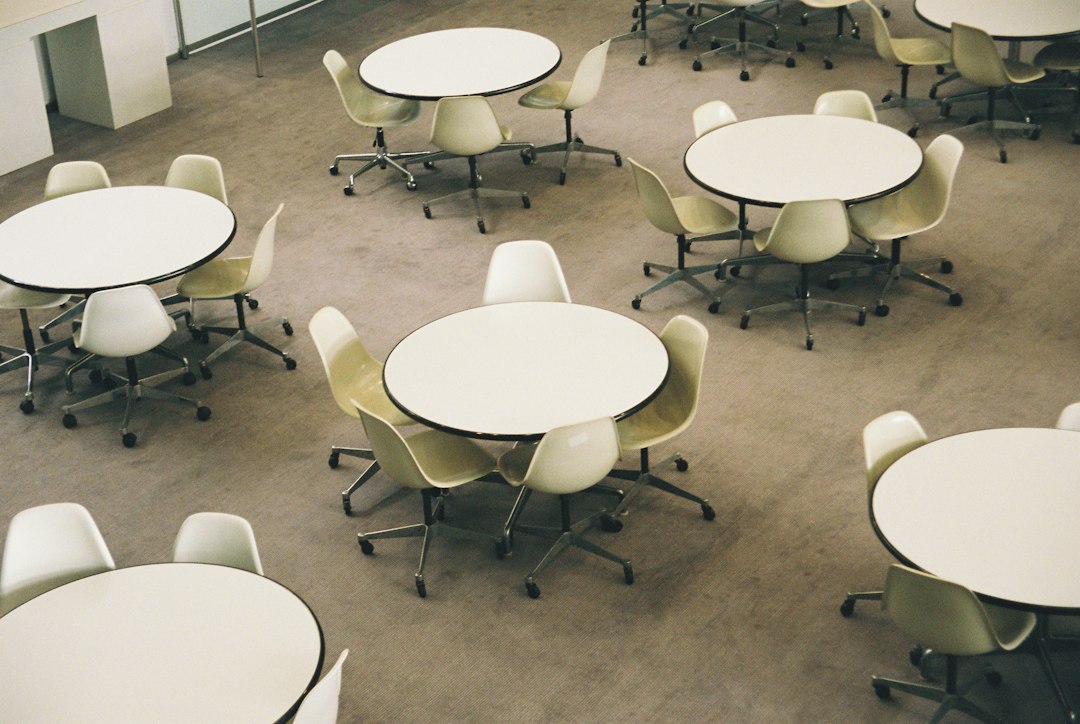 Empty meeting space with round white tables and beige swivel chairs on wheels on a gray carpet.