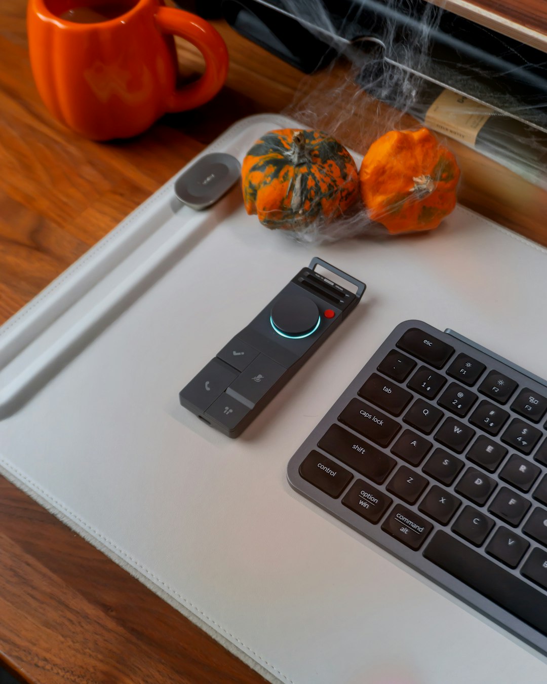 Small black media/controller device with a glowing blue ring on a white desk mat beside a keyboard, two orange pumpkins, and an orange mug with cobweb decor in autumn setup.
