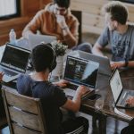 Group of people coding on laptops around a wooden table in a casual workspace.