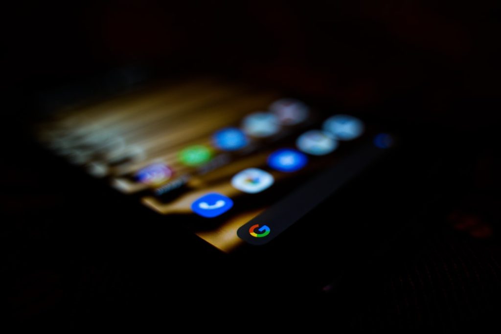 Close-up of a smartphone home screen in low light, with colorful app icons visible and the Google icon in the dock bottom-right corner.