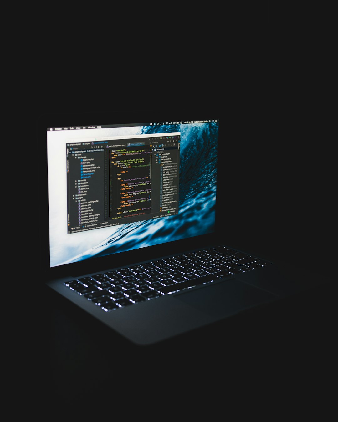 Laptop screen shows a code editor with colorful syntax highlighting against a blue ocean wallpaper in a dark setting, conveying programming working.