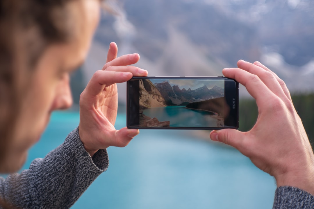 Person holding a smartphone horizontally to photograph a turquoise lake and surrounding mountains in a valley