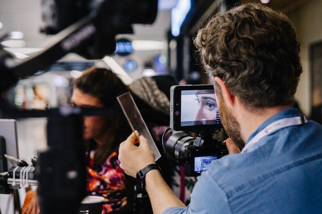 Cinematographer operating a professional camera with an external monitor, filming a subject holding a card in a busy set.