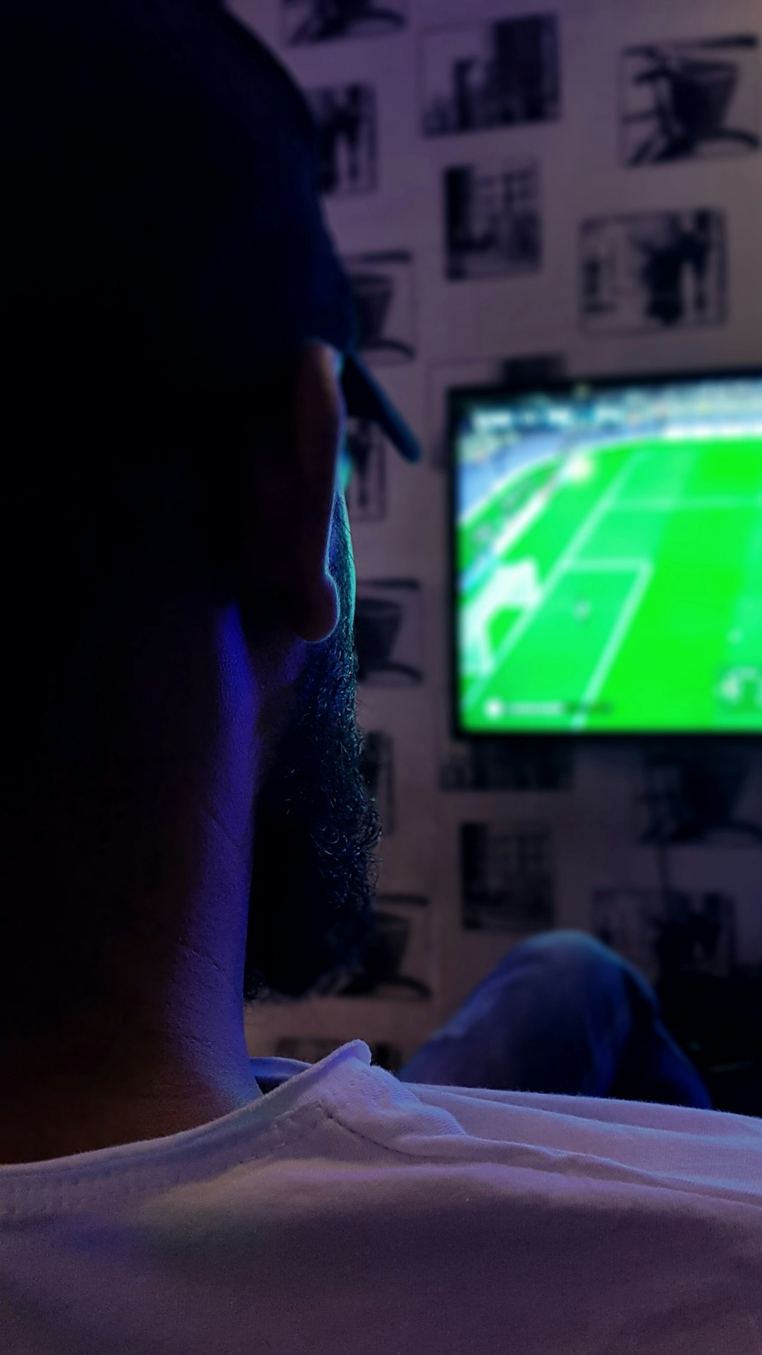 Person seen from behind watching a TV screen showing a green soccer field in a dimly lit room with wall art/posters visible in the background.