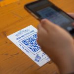 Card on a wooden table shows 'WURSTHALL' with a QR code; a person holds a phone nearby to scan it for payment/order.