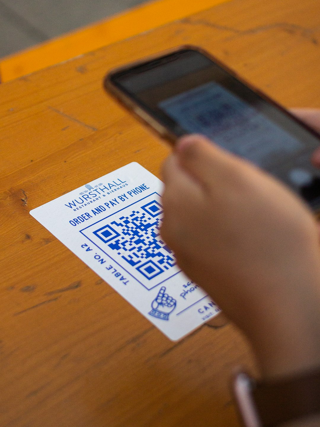 Card on a wooden table shows 'WURSTHALL' with a QR code; a person holds a phone nearby to scan it for payment/order.