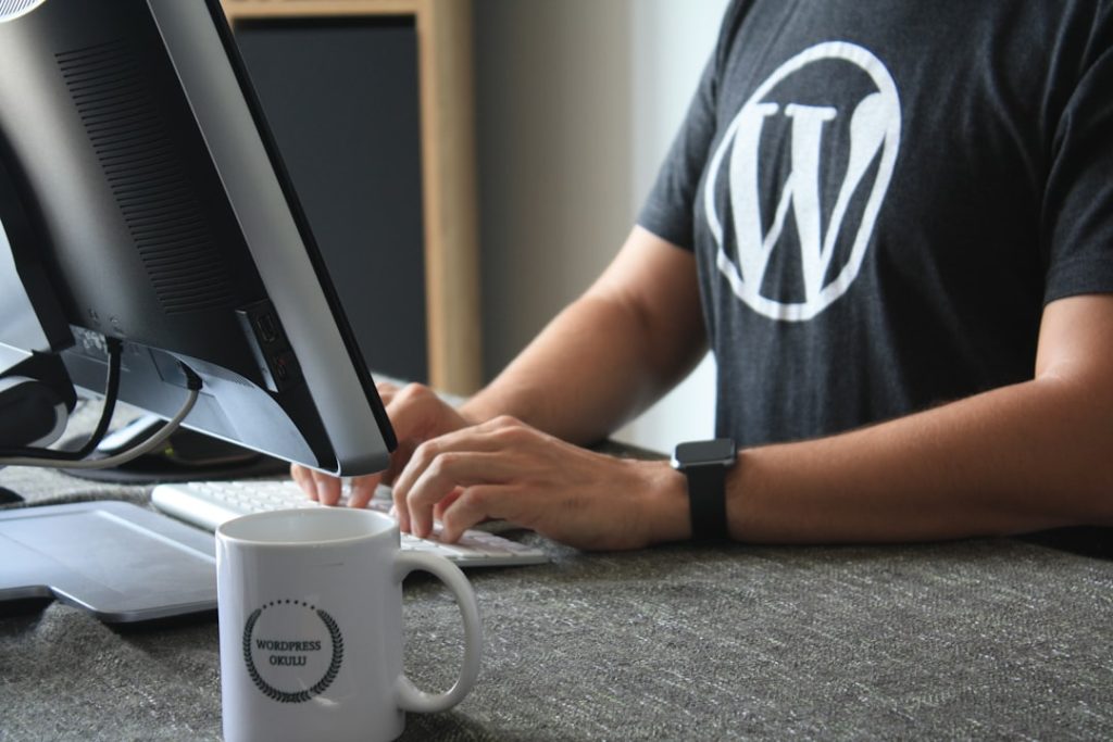Person wearing a WordPress logo shirt types at a desk, with a monitor and a coffee mug in the foreground.