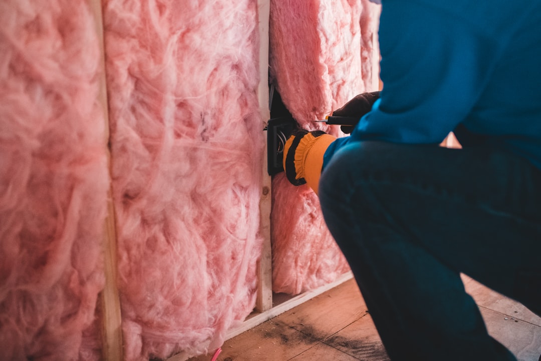 Construction worker in a blue jacket installs an electrical outlet in a wall with pink insulation.