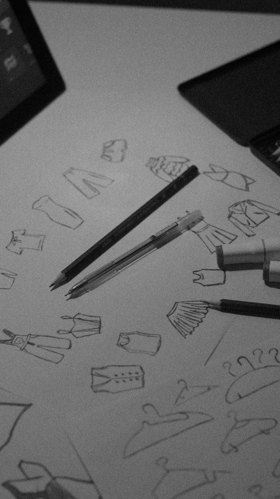 Black-and-white photo of fashion sketches on paper, with small garment drawings and two pencils nearby.
