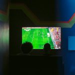 Two people sit in a dark room watching a soccer match on a bright flat-screen TV with blue neon lighting nearby.