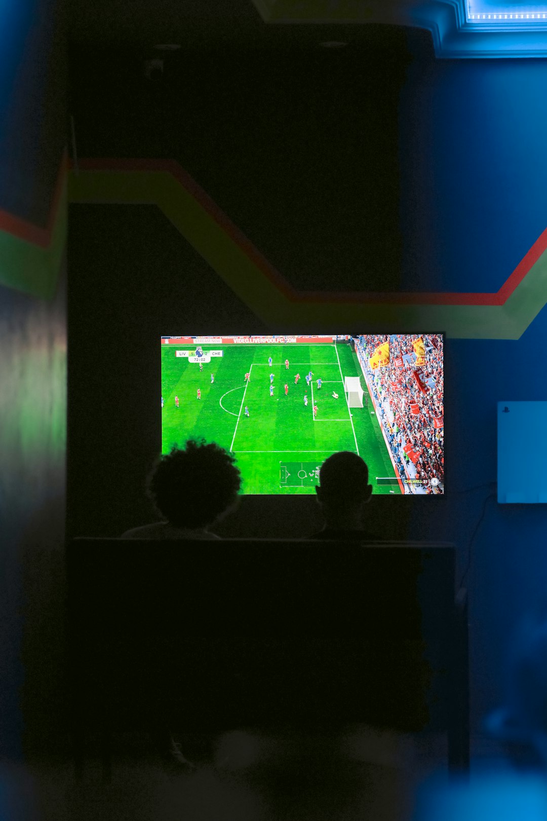 Two people sit in a dark room watching a soccer match on a bright flat-screen TV with blue neon lighting nearby.