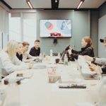 Team members sit around a conference table in a bright meeting room, watching a presentation on a wall-mounted monitor.