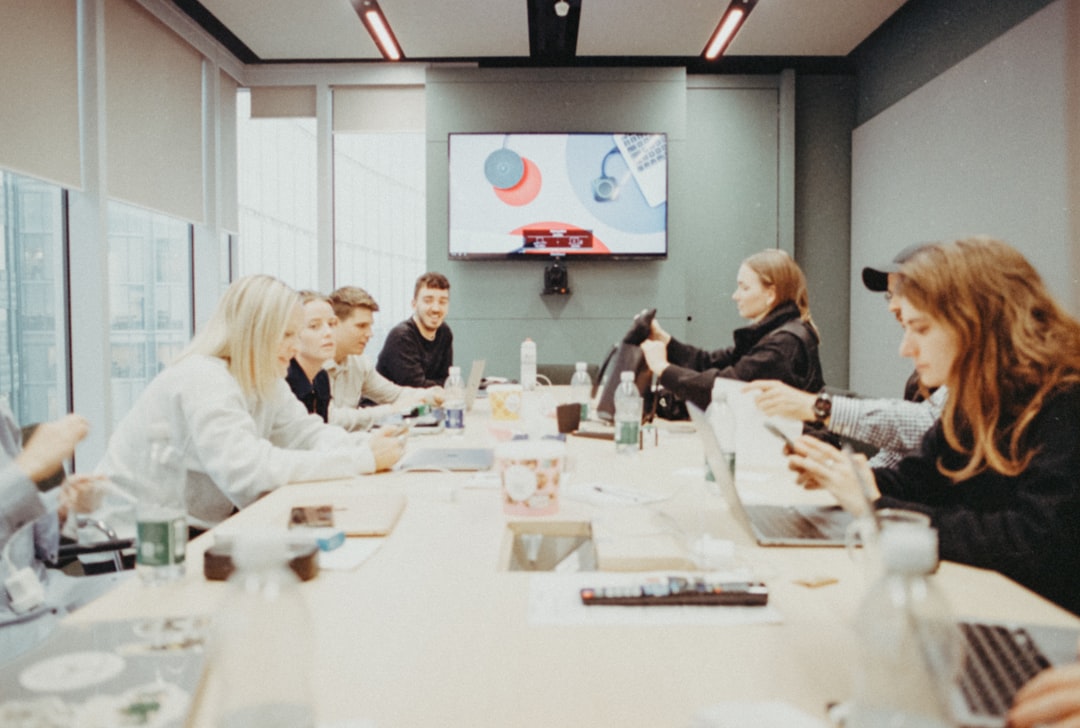 Team members sit around a conference table in a bright meeting room, watching a presentation on a wall-mounted monitor.