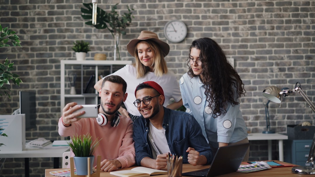 Four coworkers take a selfie around a desk in a brick-wall office with plants and a clock behind them to the left.