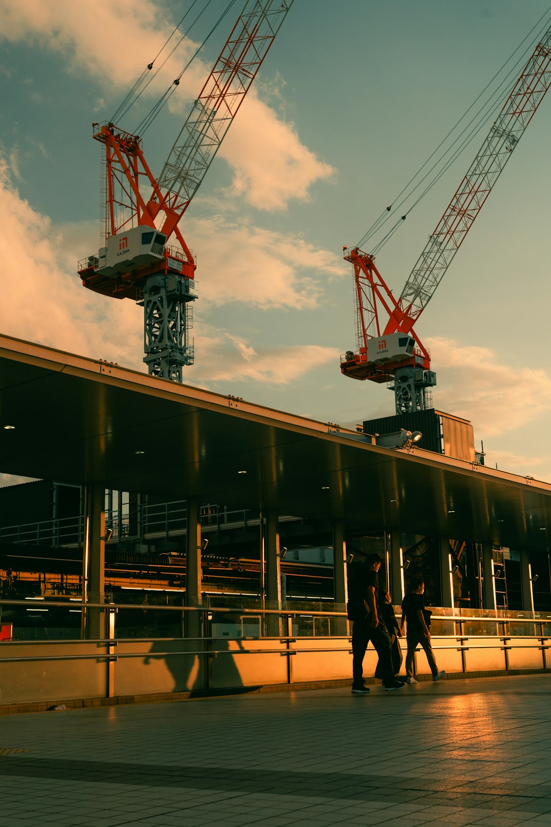 Two large red-and-white construction cranes loom over a modern building as workers walk along a covered walkway at sunset.