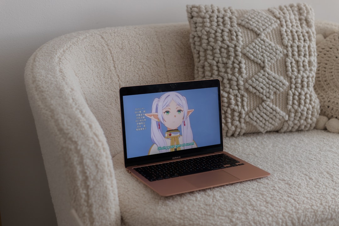 Rose-gold laptop open on a plush cream sofa with a chunky knit pillow in the background, screen displays anime character.